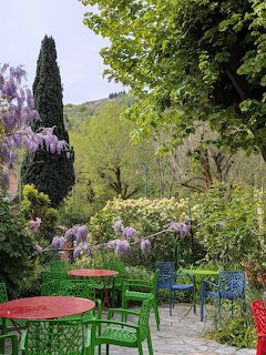 Garden terrace with flowering purple wisteria, green tables and chairs, cypress tree and mountain view