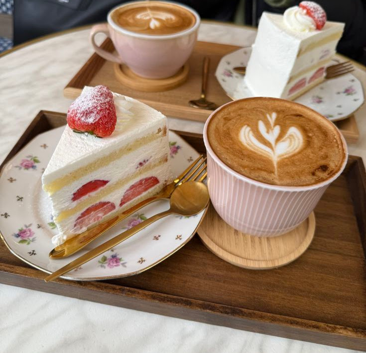 Two slices of strawberry cream cake with two lattes on wooden trays at a cafe table