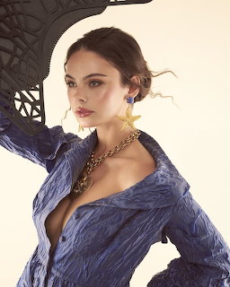 Woman in blue crinkled blazer with gold star earrings and chain necklace posing against beige background