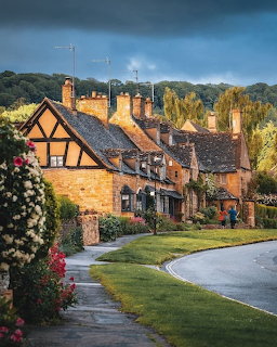 Old stone village cottages beside a curved road with flowers, lawns, and a dark cloudy sky