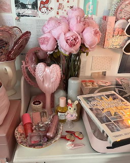 Pink peonies in a vase beside a pink angel wing hair brush, cosmetics, and accessories on a white vanity