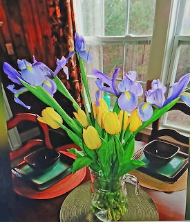 Glass vase of yellow tulips and purple irises on a dining table near a window