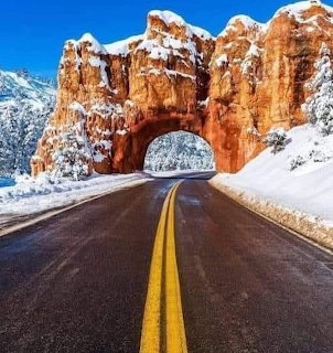 Road with yellow lines passing through a snow-covered red rock tunnel arch under blue sky