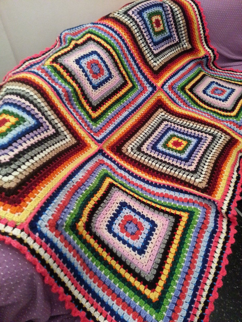 Multicolored crochet blanket with rainbow granny square pattern draped on purple surface