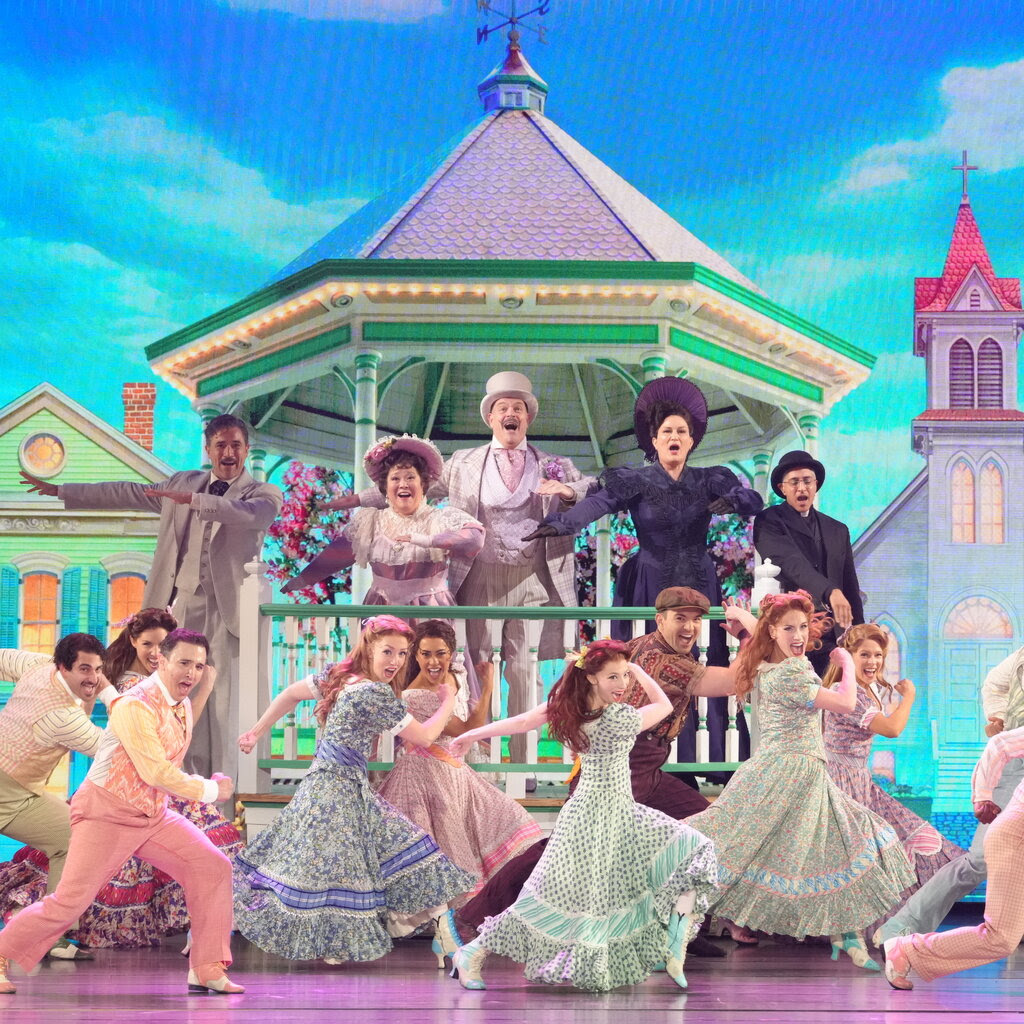 Colorful ensemble of performers posing in front of a gazebo and pastel buildings