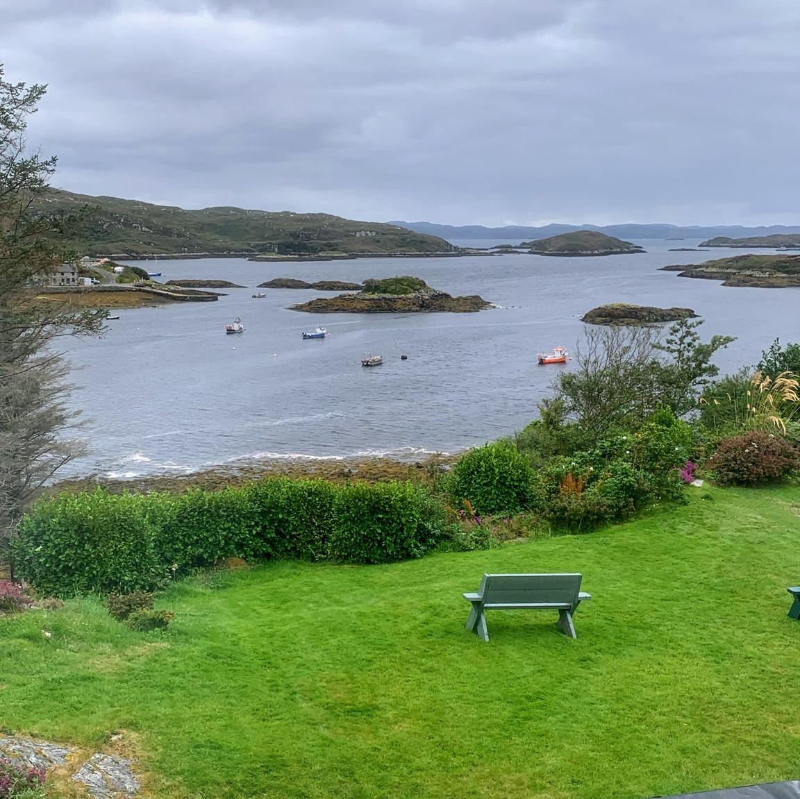 Scenic view of Badcaul Bay with islands, green lawn, bench, and hills under cloudy sky
