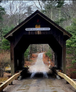 Wooden covered bridge with dark timber frame on a wet gravel road surrounded by leafless trees