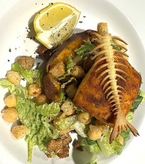 Plated salmon fillet on Caesar salad with croutons, chickpeas, fish bone garnish, and lemon wedge