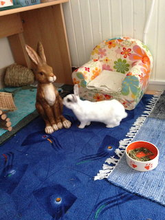 White rabbit on a blue rug beside a wooden bunny figure, floral chair, and food bowl indoors