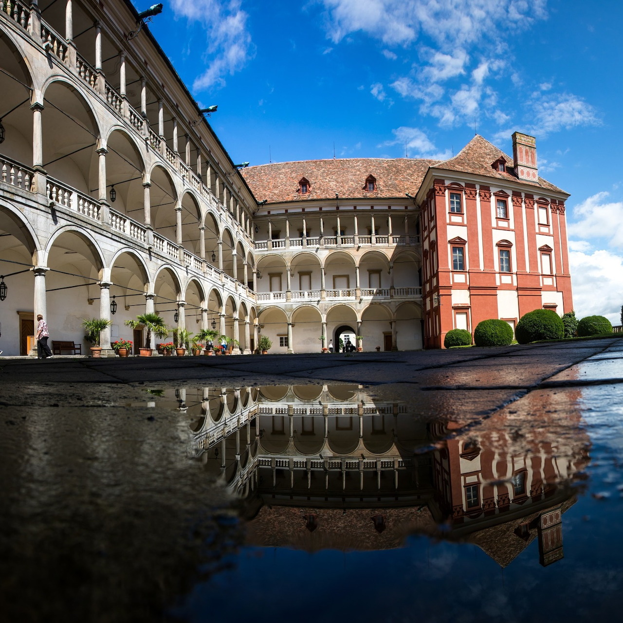 Castle courtyard with white arched galleries and coral tower reflected in water puddle