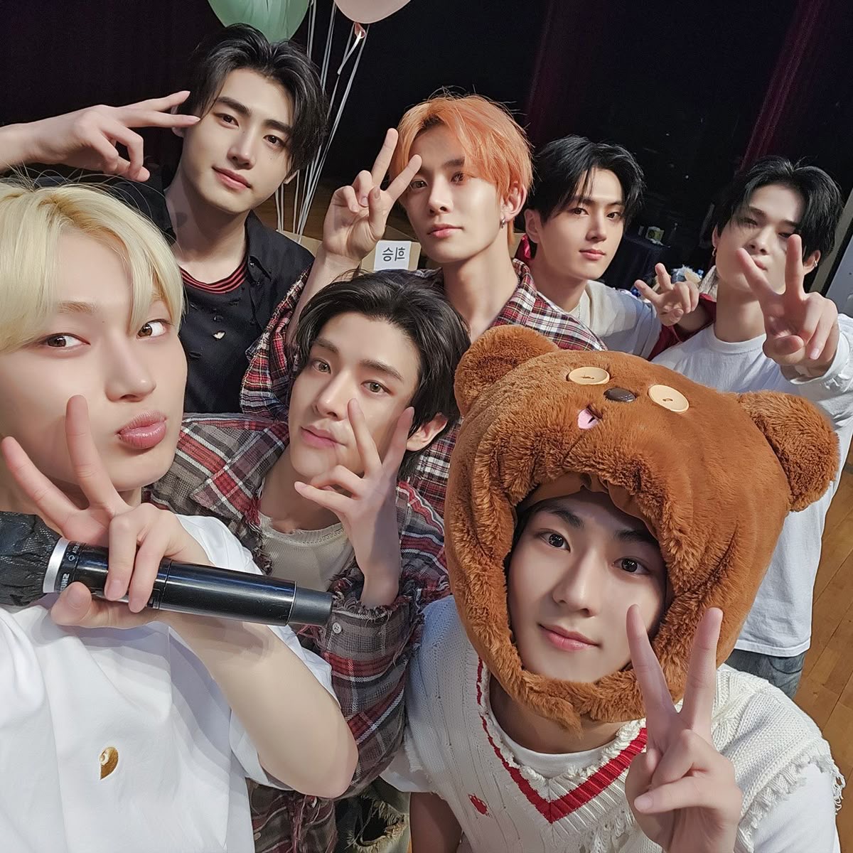 Group of seven young men in casual clothing posing playfully together, one wearing a brown bear hood