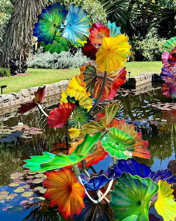 Colorful blown glass flower sculptures in red, blue, yellow, and green above a pond with lily pads