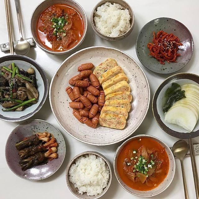 Top-down Korean meal with rice bowls, soup, omelet, sausages, kimchi, vegetables, and side dishes