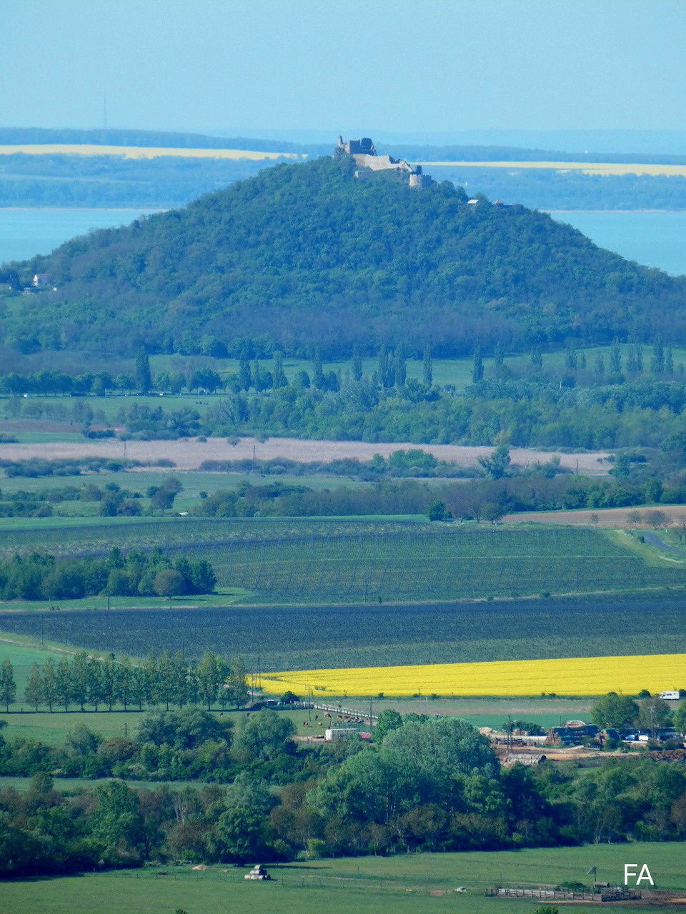 Green hill with castle ruins surrounded by farmland, yellow rapeseed fields, and a distant lake