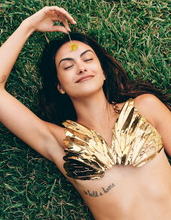 Woman lying on green grass wearing a gold metallic top with eyes closed and a flower on her forehead