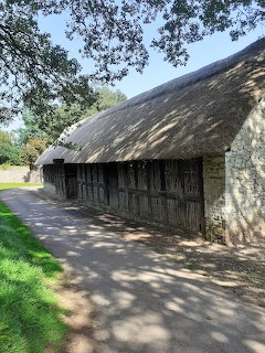Stone barn with thatched roof and wooden doors along a paved path under oak trees