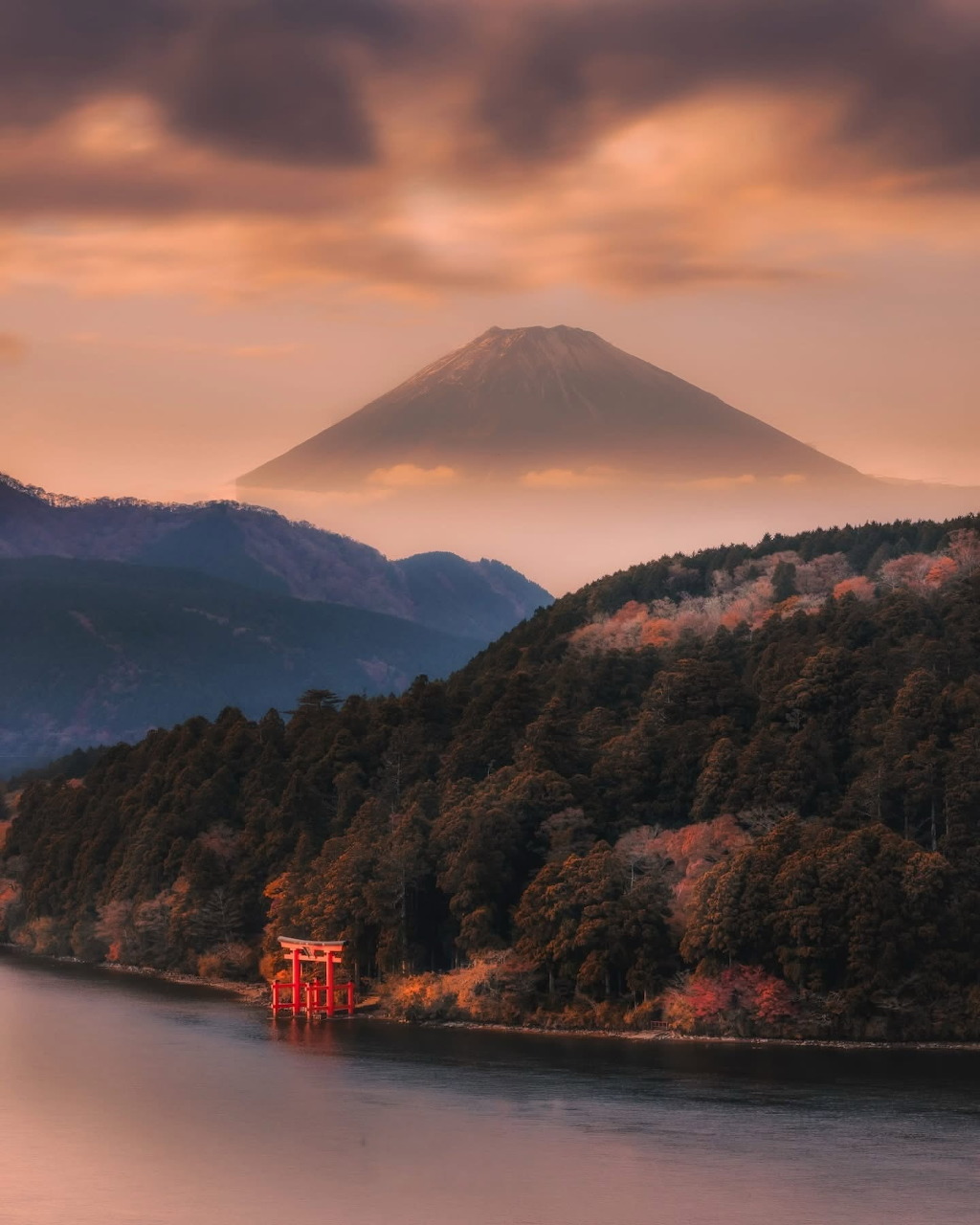Mount Fuji silhouette at sunset with red torii gate on lake shore and forested mountains in foreground