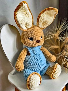 Crocheted amigurumi rabbit toy in tan and blue overalls sitting on a white chair