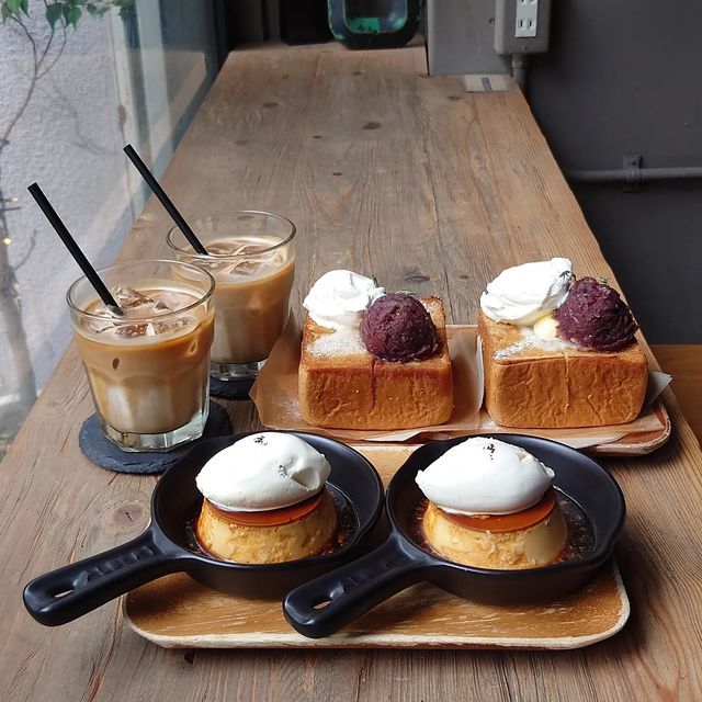 Cafe table with iced coffee, purin desserts in black skillets, and toast with toppings on wooden serving boards