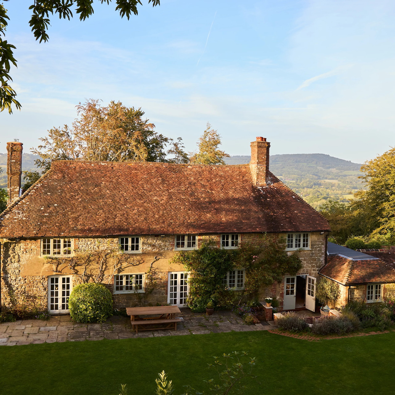 Stone manor house with red tile roof and lattice windows on green lawn with hills behind