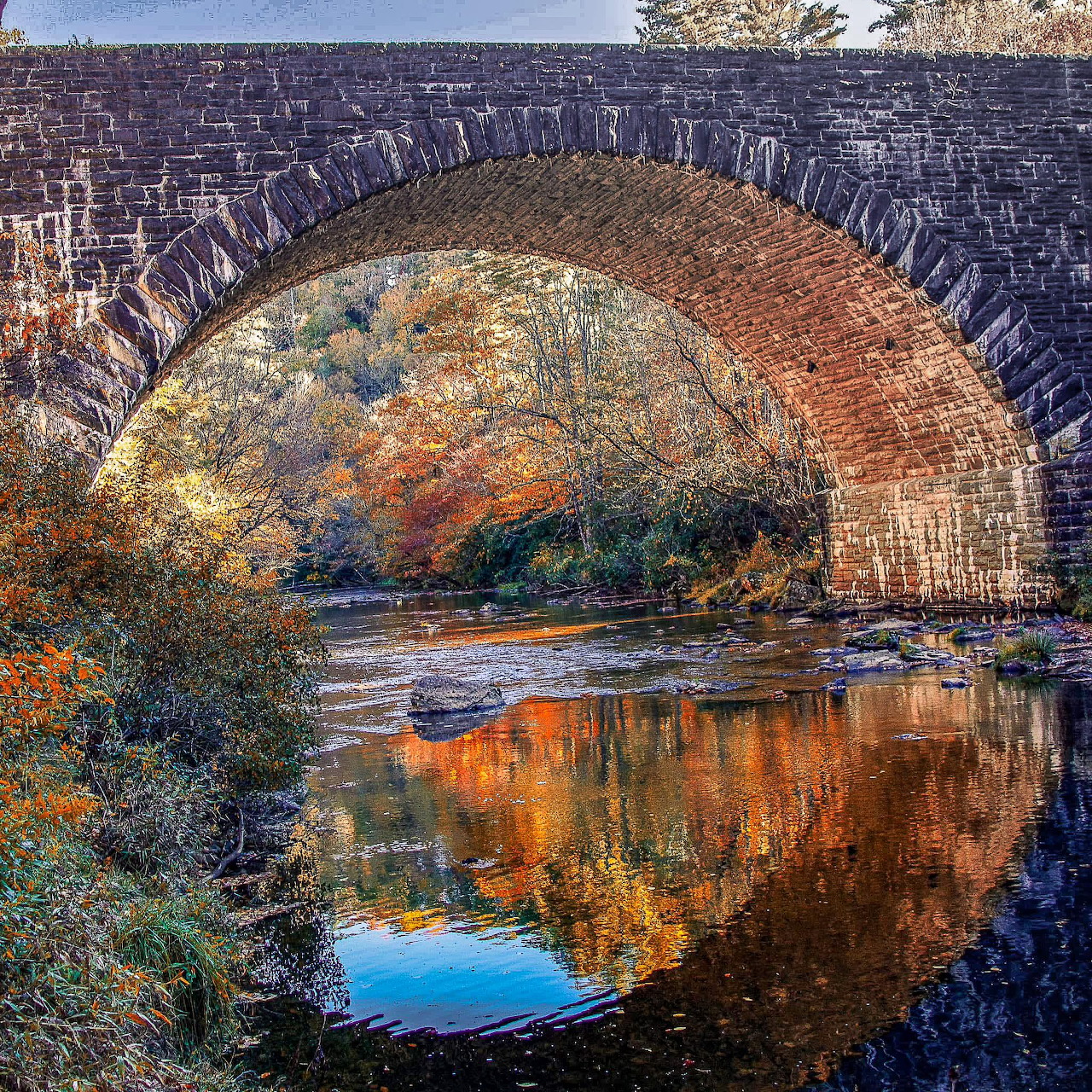 Stone arch bridge over a river with autumn foliage reflected in the water below