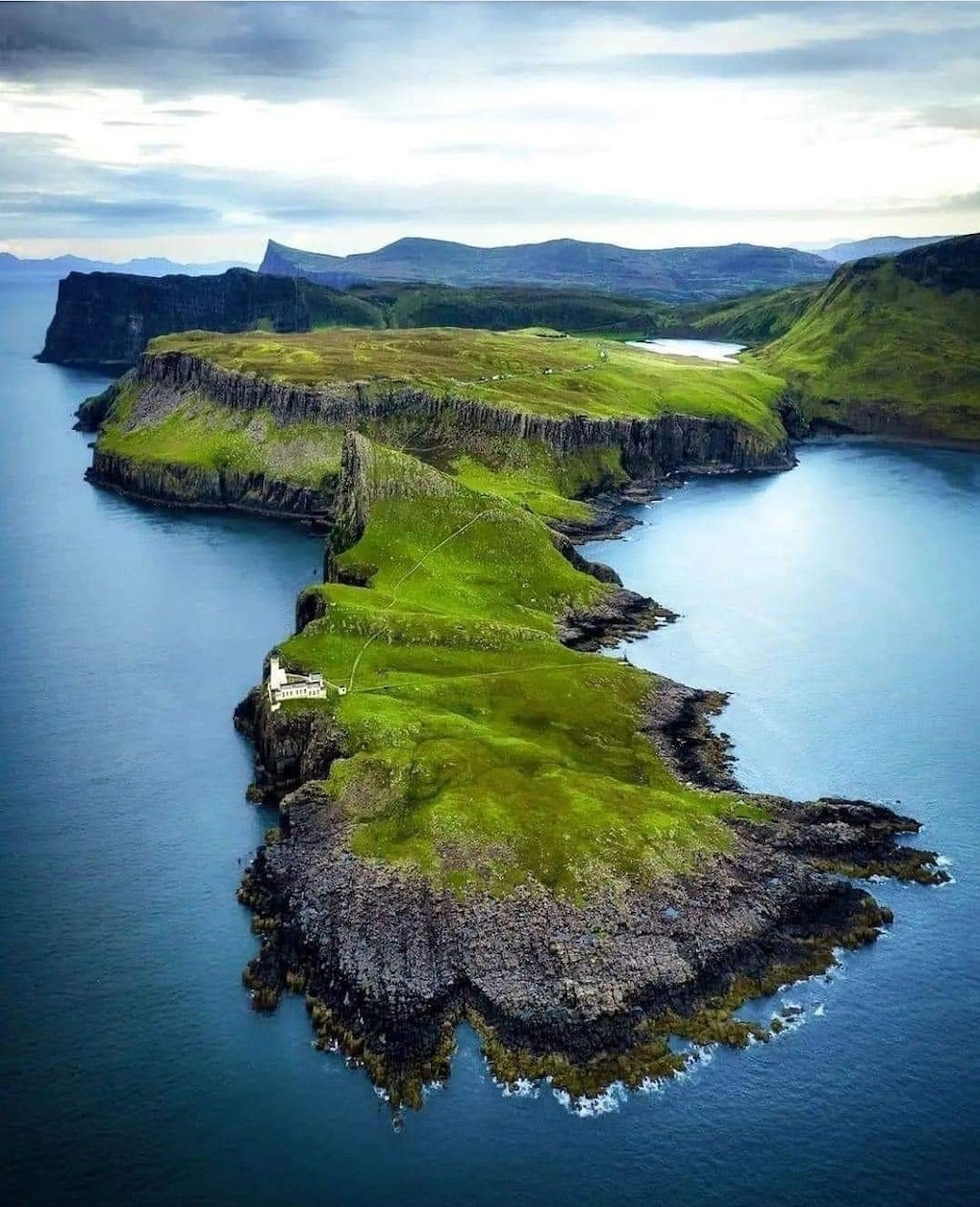 Aerial photo of green cliffside peninsula extending into blue ocean waters with mountains in background