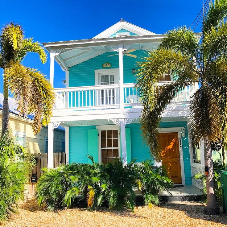 Bright turquoise house with white porch, balcony, and palm trees in a tropical setting