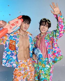 Two men in bright floral jackets waving happily against blue background