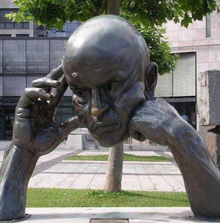 Large bronze statue of a pensive man resting his head on both hands, outdoors in a city plaza