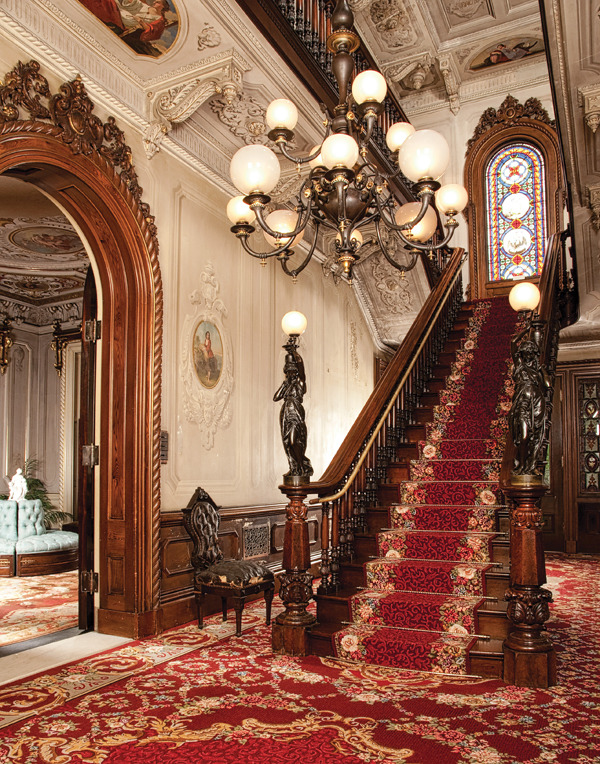 Grand curved staircase with red carpet in ornate Victorian mansion interior with chandelier and stained glass