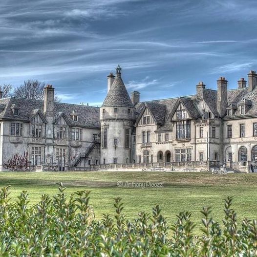 Large historic stone mansion with a central turret, green lawn, and blue sky
