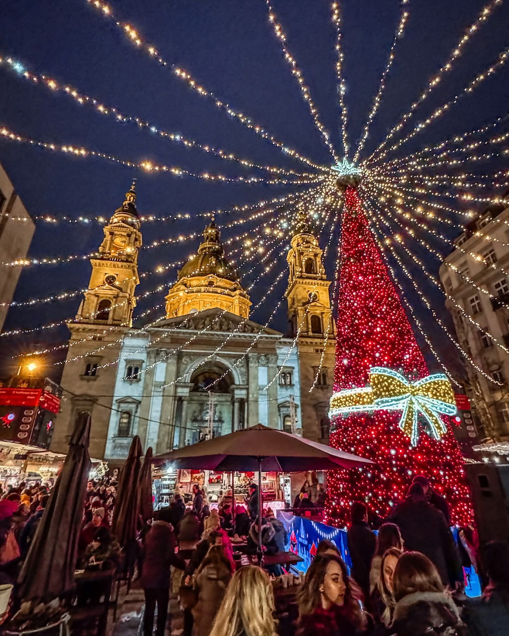 Christmas market at night with large illuminated tree and historic basilica, festive lights radiating overhead