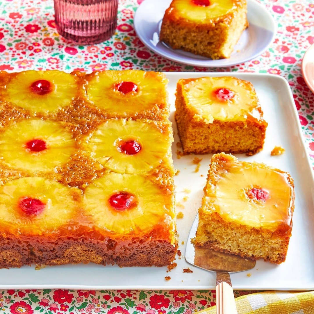 Pineapple upside down cake with pineapple rings and cheries on a white platter, sliced pieces beside it