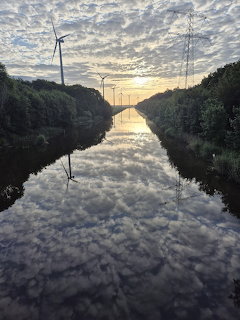 Canal reflecting clouds and sunset light between tree-lined banks with wind turbines and power lines