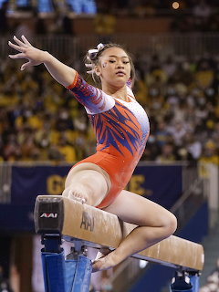 Female gymnast mid-routine on balance beam with arms outstretched in orange and blue competition leotard