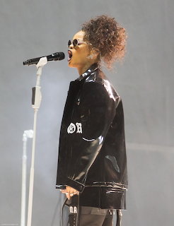 Rihanna singing into a microphone on stage wearing a black leather jacket and round sunglasses