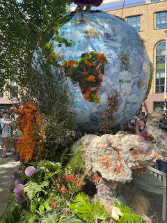 Spherical sculpture covered in flowers and plants resembling Earth, surrounded by garden plants and rocks
