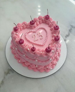 Pink heart-shaped frosted cake topped with cherries on a white cake board