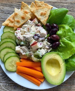 White plate with chicken salad, cucumbers, carrots, avocado, olives, lettuce, and crackers on wood table