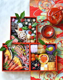 Japanese bento box with sushi, sashimi, prawns, and vegetables arranged in compartments with tea and decorative fabric
