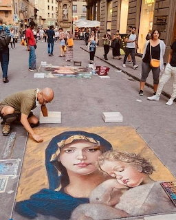 Street artist drawing a large chalk Madonna and Child painting on a city sidewalk with pedestrians watching