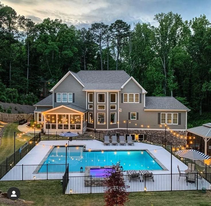 Large gray house behind a fenced swimming pool with patio lights and trees at dusk