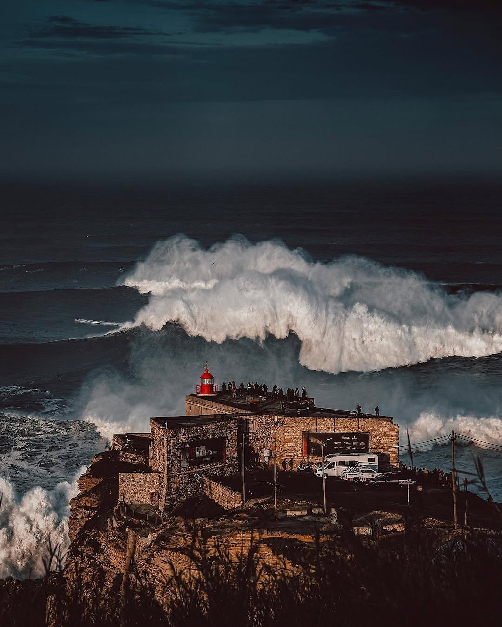 Enormous wave crashing behind a red lighthouse and stone building on a rocky cliff by the sea