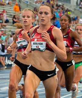Female runners competing in a track race wearing athletic uniforms with spectators in background