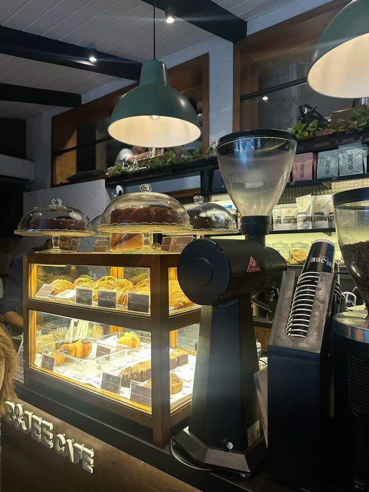 Café counter with illuminated glass pastry display case, coffee grinder, and green pendant lights overhead.