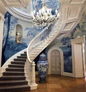 Elegant white curved staircase with chandelier in room with blue floral wallpaper and ornate ceiling