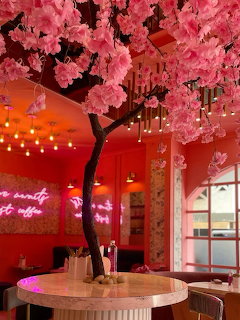 Pink cafe interior with artificial cherry blossom tree, neon signs, and round marble table