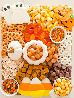 Halloween snack board with ghost cookies, candy corn, pretzels, chocolate, and orange treats arranged on white background