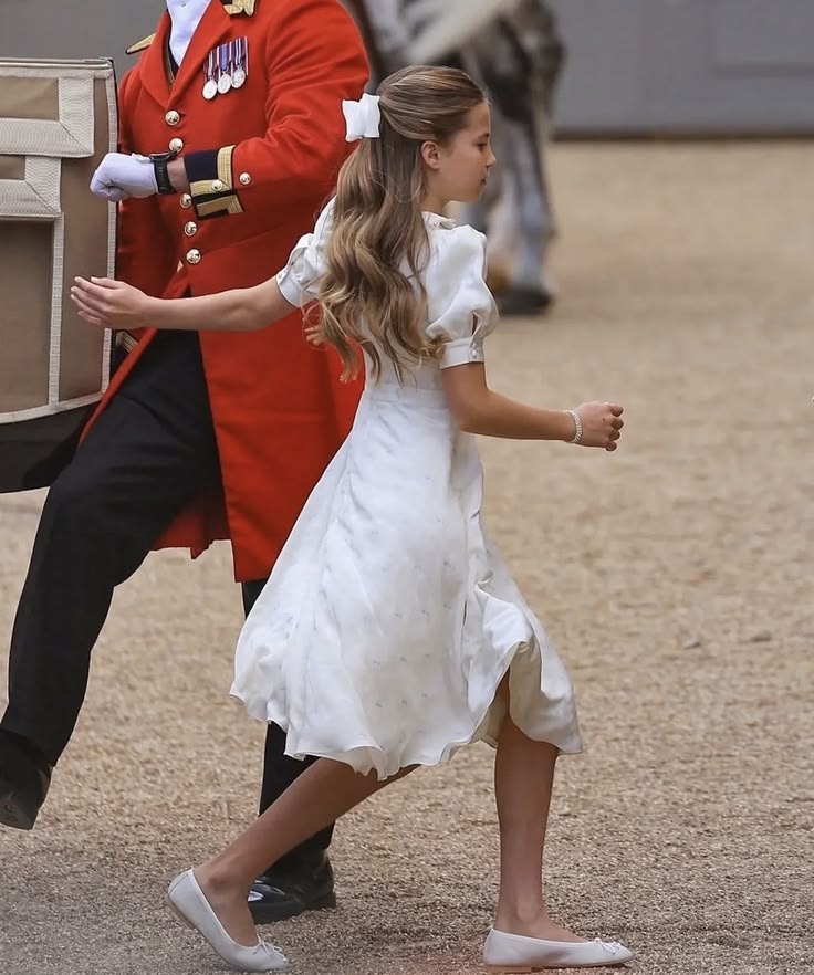 Young girl in white dress with hair bow walking past a guard in red uniform at a royal event