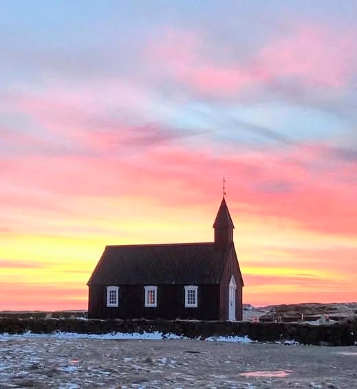 Small black church on snowy ground under a colorful pink and orange sunrise sky
